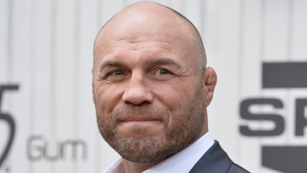 Randy Couture in abito grigio scuro e camicia bianca, guardando fuori di lato con un piccolo sorriso