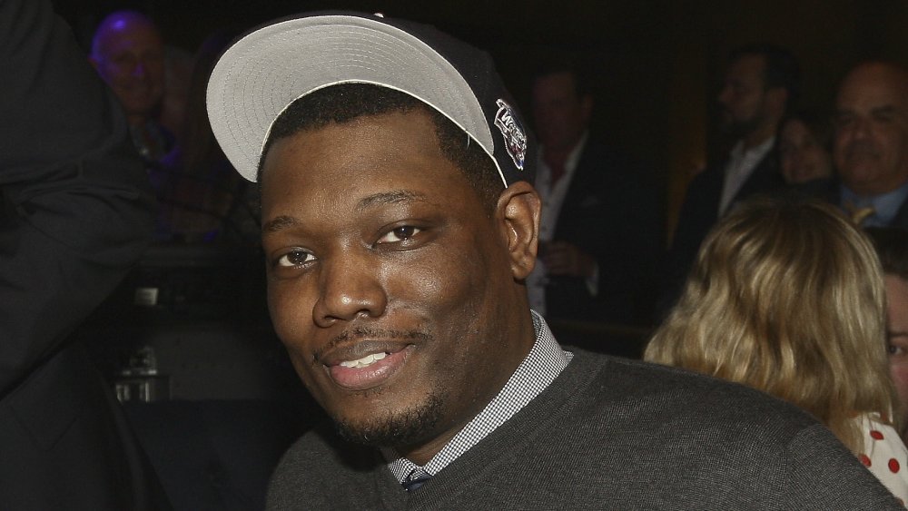 Michael Che in un maglione grigio e cappello nero, in posa con un piccolo sorriso