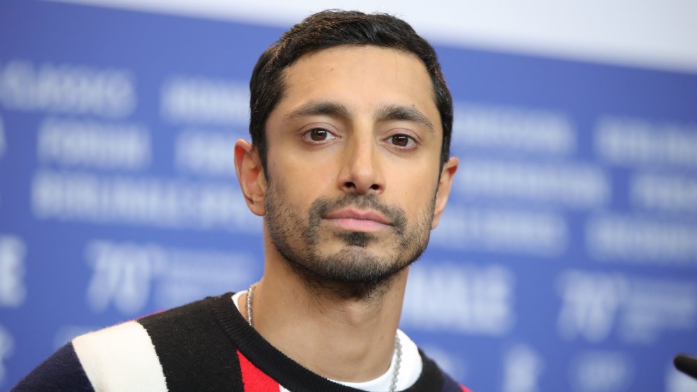 Riz Ahmed in un maglione nero, rosso e bianco, in posa durante un'intervista con un'espressione neutra