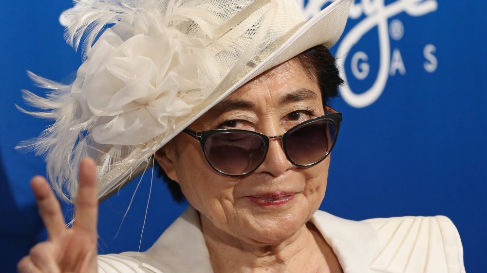 Yoko Ono in un blazer bianco, cappello e occhiali da sole, sorridendo e dando il segno di pace