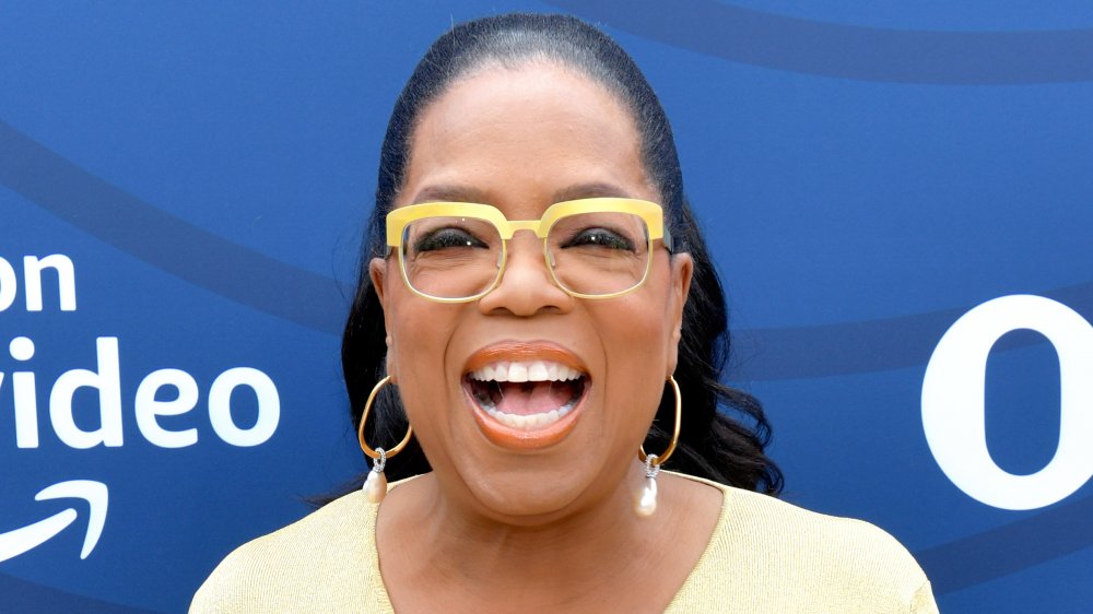 Oprah Winfrey in abito giallo e occhiali, ridendo mentre guarda dritto la fotocamera