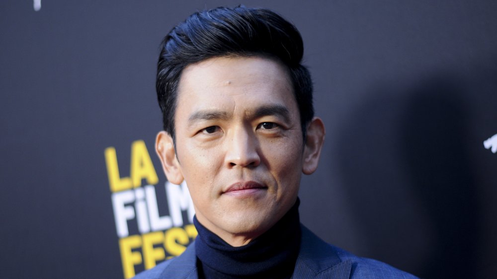 John Cho in un blazer blu e dolcevita, in posa con un'espressione seria