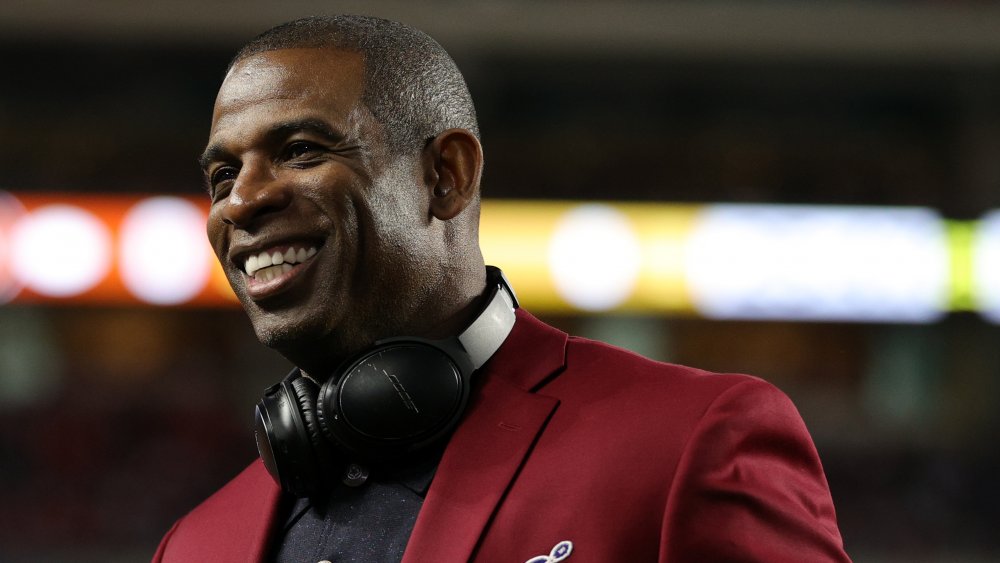 Deion Sanders sorridente, mentre guarda di lato, in un blazer marrone e polo nera