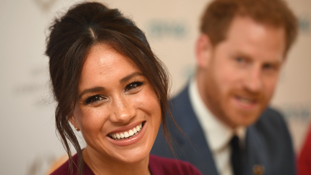 Meghan Markle sorride con il principe Harry sullo sfondo