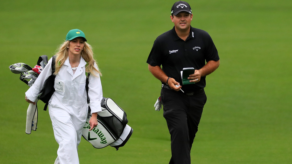 Justine Reed, che lavora come caddy, vestita di bianco, indossa un odio, tiene in mano mazze da golf;  Patrick Reed, vestito di nero e cappello, sul campo da golf