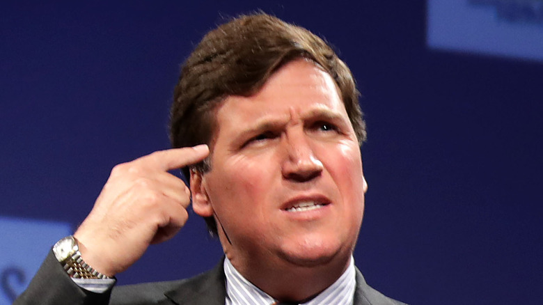 Tucker Carlson con un dito contro la testa, strabico