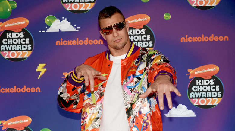 Rob Gronkowski indossa una giacca arancione con fantasia neon, una maglietta bianca e occhiali da sole scuri mentre punta verso la telecamera