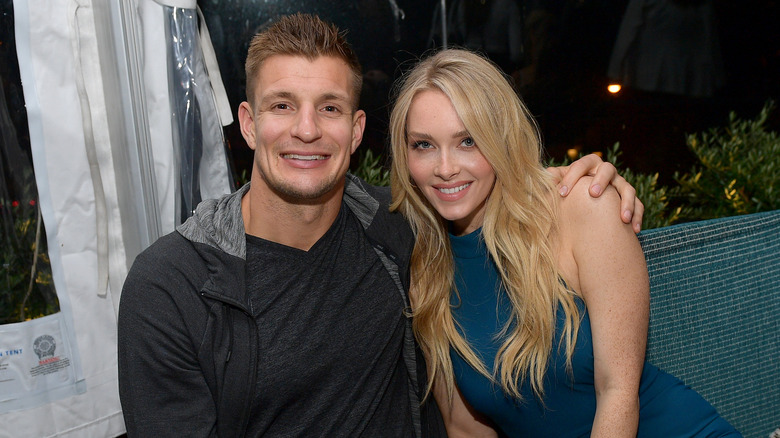 Rob Gronkowski indossa una giacca grigia e una maglietta grigia con Camille Kostek che indossa un abito senza maniche a collo alto color verde acqua