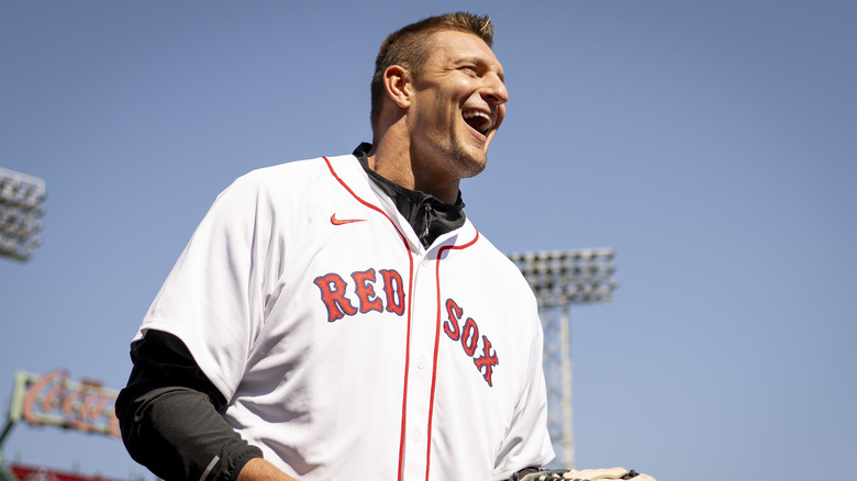 Rob Gronkowski sorride mentre indossa una maglia da baseball bianca dei Boston Red Sox e una camicia nera a maniche lunghe