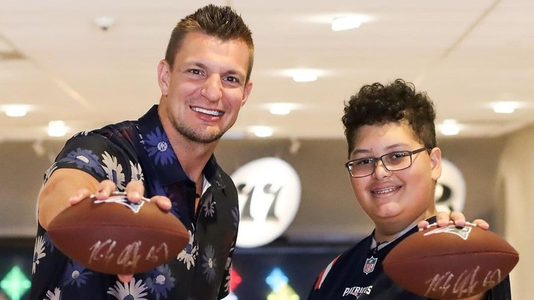 Rob Gronkowski sorride tenendo in mano un pallone da football con un bambino che indossa occhiali e una maglia da football dei Patriots