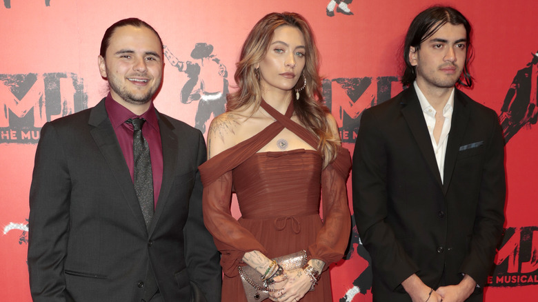 Prince Jackson indossa una giacca nera, una camicia rossa e una cravatta nera; con Paris Jackson indossa un abito incrociato color ruggine; con Bigi Jackson indossa una giacca nera e una camicia bianca