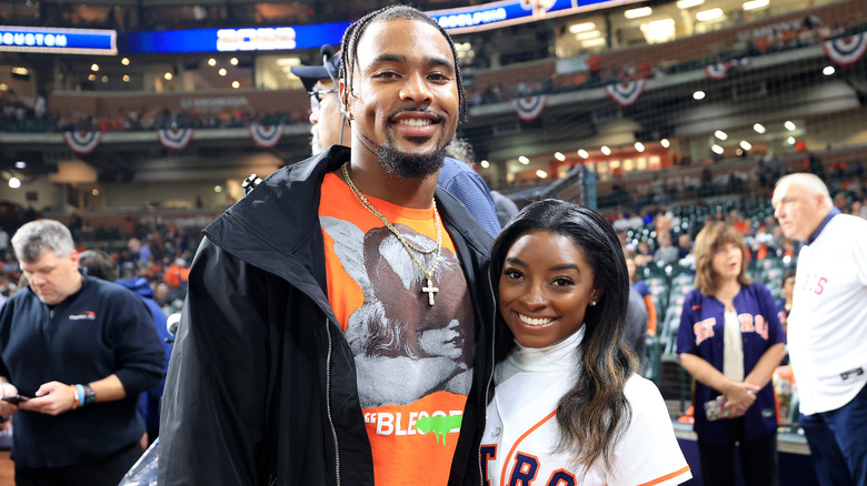 Jonathan Owens indossa una maglietta grafica arancione e una giacca nera con Simone Biles che indossa una maglia da baseball bianca degli Houston Astros e un dolcevita bianco