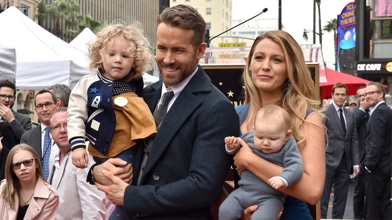 Ryan Reynolds indossa un abito nero e cravatta con Blake Lively in un abito blu mentre entrambi tengono in braccio i loro figli