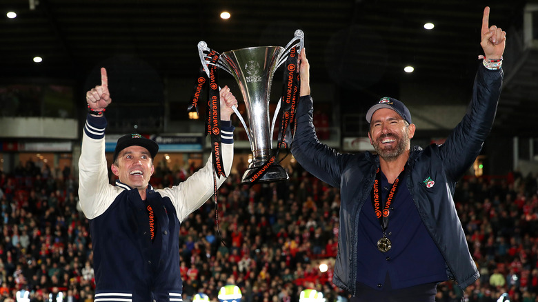 Rob McElhenny e Ryan Reynolds indossano l'attrezzatura del Wrexham AFC mentre tengono in mano un trofeo