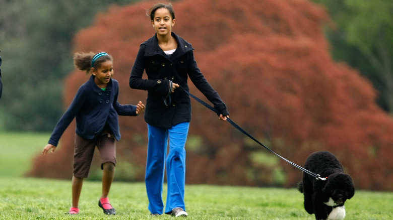 Sasha Obama indossa una giacca blu e pantaloncini marroni con Malia Obama che indossa un cappotto nero, jeans blu e tiene un cane al guinzaglio