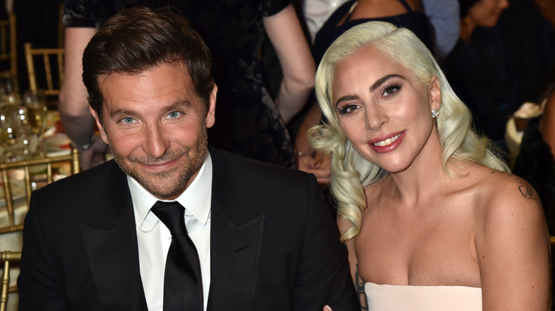 Bradley Cooper in abito nero e cravatta seduto con Lady Gaga in un abito bianco senza spalline