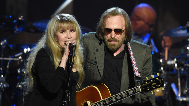 Stevie Nicks indossa una giacca e un vestito neri mentre canta con Tom Petty che indossa una giacca grigio-verde e una camicia nera