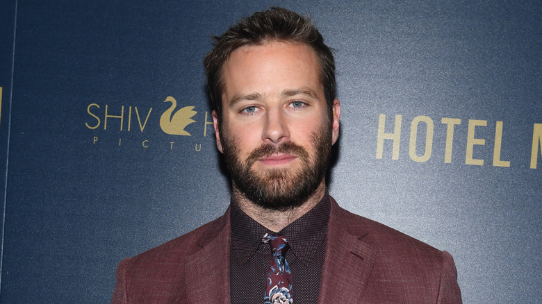 Armie Hammer indossa una giacca marrone e una camicia nera con fantasia e una cravatta con motivi cachemire