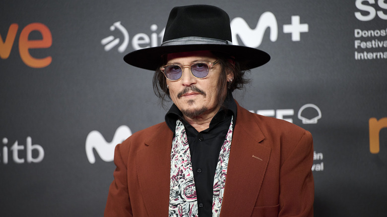 Johnny Depp indossa una giacca arancione bruciato, una sciarpa con motivi cachemire, una camicia nera, un cappello nero e occhiali da sole colorati