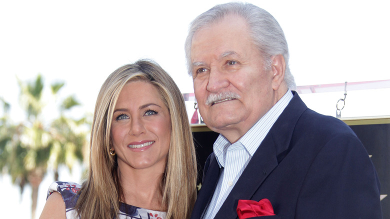 Jennifer Aniston indossa un abito floreale e John Aniston indossa una giacca blu e una camicia con colletto a righe bianche